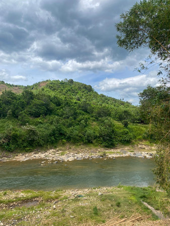 Tropical river flowing along rocky shore with lush green hillside and dramatic overcast sky.
