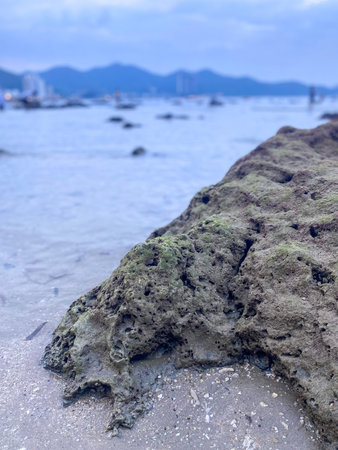 Large coastal rock formation on sandy beach with calm ocean water and distant mountains.