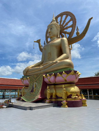 A monumental golden Buddha statue seated on a decorated pedestal at a Buddhist temple in Thailand. Behind the statue rises a large Dharma wheel symbolizing Buddhist teachings and enlightenment. The temple complex features ornate traditional architecture, golden decorative elements and open courtyards under a bright tropical sky. Such spiritual landmarks attract pilgrims and travelers exploring Thai culture, religion and Southeast Asian heritage.の写真素材