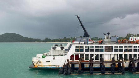Passenger ferry docked near tropical island coastline with cloudy sky and turquoise sea in Thailand.の写真素材