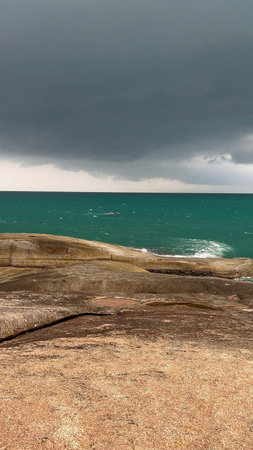 Turquoise ocean waves meeting a granite rocky shoreline beneath heavy storm clouds in a coastal...