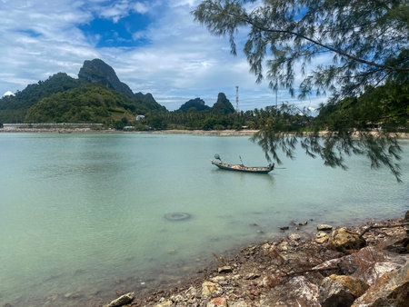 Traditional longtail boat anchored near rocky shoreline with tropical hills and calm sea.