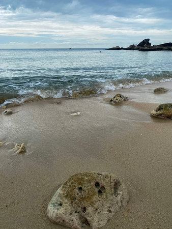Small ocean waves rolling onto sandy beach with stones and cloudy sky.の写真素材