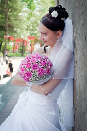 the beautiful young bride stands near a brown stone wall with a pink bouquet in handsの写真素材