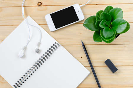Top view of Blank white Notebook and black pencil on wooden table with smart phone and small flowerpotの写真素材