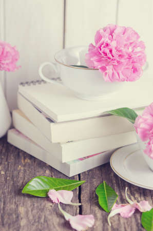 Pink pastel carnation flowers in white tea cup and vase on pile of books on wooden tableの写真素材