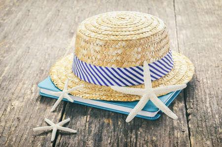 Beach concept - Straw Hat with white and blue striped on wooden table with book and starfishes with sunlight and vintage toneの写真素材