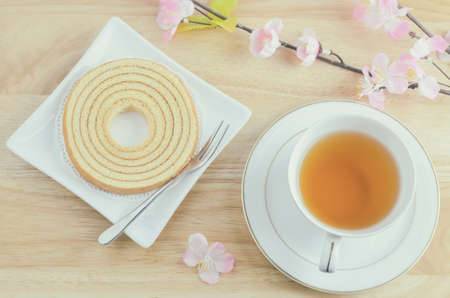 Baumkuchen sweet layered cake at white dish with cup of hot tea and artificial pink sakura flower on wooden table with vintage toneの写真素材
