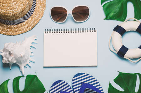 Blank white notebook paper with straw hat, Seashell, sunglasses, blue sandal, lifebuoy and green leaves on blue wooden background with vintage tone - Summer backgroundの写真素材