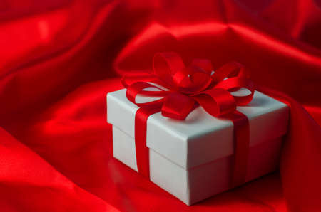 Close up of white gift box with red ribbon bow on red satin textile backgroundの写真素材