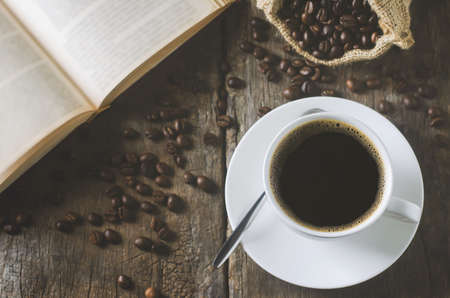 White cup of black coffee on wooden table with coffee beans and open book with vintage and viggnete toneの写真素材