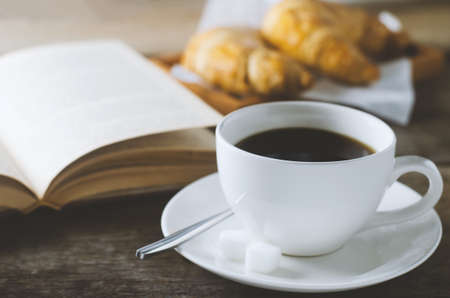 Close up of black coffee with book and croissant on wooden table with vintage and vignette toneの写真素材