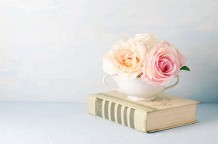 Artificial pink rose flowers in white cup with old book on blue background with vintage toneの写真素材