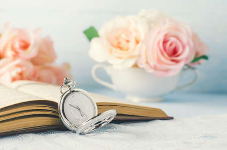 Close up of antique silver pocket watch and opened book with rose flowers on white and blue background with vintage toneの写真素材