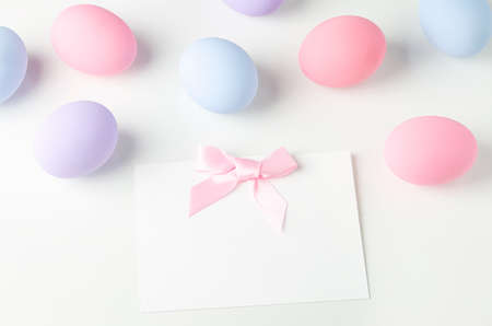 Blank white greeting card with pink ribbon bow and pastel easter eggs on white tableの写真素材