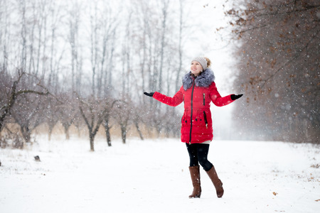 Happy beautiful girl in knitted hat and red winter coat, outdoors in park, rejoicing at snowfall, full lengthの写真素材