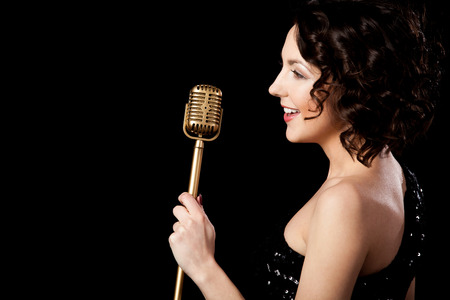 Elegant young female singer in shiny black evening outfit singing with smile, holding golden vintage microphone, live performance, concert, copy spaceの写真素材
