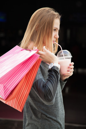 Attractive teenage girl out for having fun, shopping, walking in store with colorful paper bags, drinking strawberry milkshake, enjoying herselfの写真素材