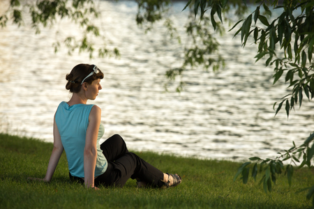 Young female sitting on the grass on the river bank, waiting, looking into the distance, dreaming, resting, relaxing, on the break in warm calm weatherの写真素材