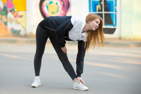 Smiling runner girl with earphones in sportswear doing stretching exercises before morning jogging on the sunny street in front of wall with bright colored graffiti, preparing for runningの写真素材