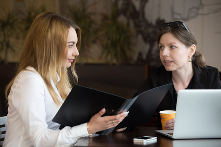 Two young caucasian women sitting at the table in cafe beside laptop, having discussion, blond girl holding open document folder, listeningの写真素材