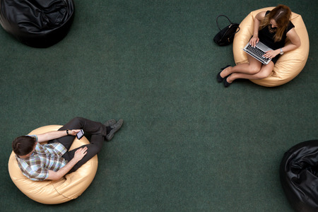 Two young people, man and woman, sitting on bean bags, using electronic devices in public wifi area, working on laptop, typing, using smartphone, texting message, top viewの写真素材