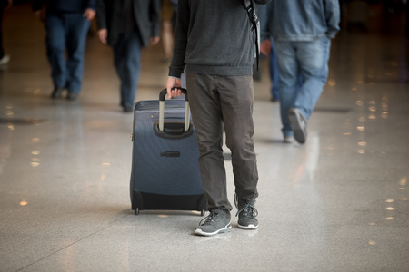 Young man with suitcase walking in modern airport terminal with crowd on background, wearing casual style clothes, close upの写真素材