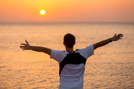 Young man standing at the seashore with raised arms feeling happy, watching colorful sunrise or sunset sky in carefree pose, enjoying freedom, beautiful sea view and fresh airの写真素材