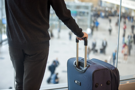 Young handsome man in 20s waiting for flight, standing in modern airport terminal with crowd on background, holding handle of luggage bag, wearing casual clothes, rear view, close upの写真素材