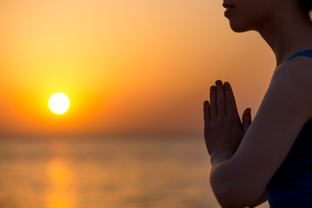 Profile of serene young woman relaxing on the beach, meditating with hands in Namaste gesture at sunset or sunrise, close upの写真素材