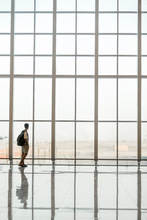 Minimalistic shot of full length silhouette of young handsome man with backpack waiting for flight, standing in modern airport terminal building in front of giant window, back viewの写真素材