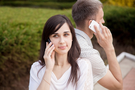 Young couple sitting back to back on park bench holding cell phones in hands, talking on mobile...