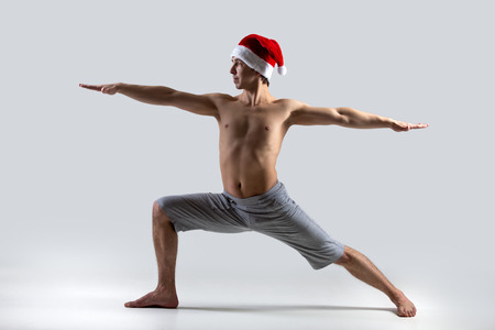 Sporty muscular young man in red Santa Claus Christmas hat working out, yoga, pilates, fitness training, doing lunge exercise, Warrior II posture, Virabhadrasana 2の写真素材