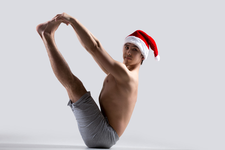 Sporty young man in red Santa Claus Christmas hat doing yoga or pilates exercises, sitting in Ubhaya Padangusthasana Both Feet Big Toe Pose or Balancing Stick Poseの写真素材