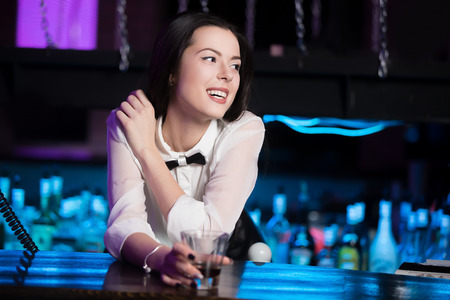 Beautiful cheerful smiling brunette girl in white shirt and black bow tie at nightclub bar, holding glass with drinkの写真素材