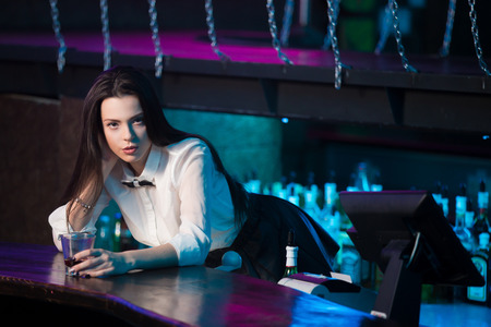 Beautiful sensual brunette bartender girl in white shirt and black bow tie working at nightclub bar, offering glass with drinkの写真素材