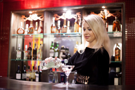 Beautiful smiling blond bartender girl serving alcohol drink at bar counter, holding bottle in hand, pouring drink in cocktail glassの写真素材