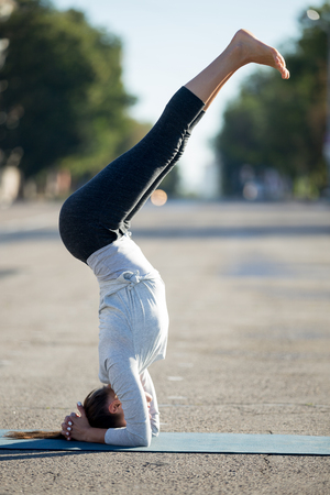 Yoga in the city: beautiful young sporty woman doing supported headstand posture, variation of salamba sirsasana in the street on summer day, full length, side viewの写真素材