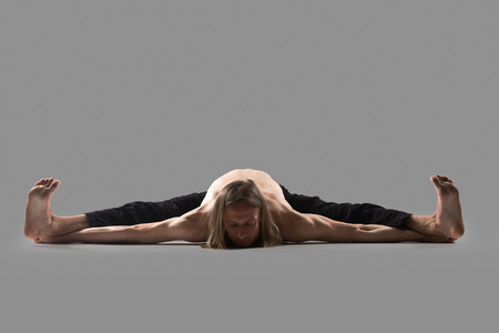 Sporty young man in black trousers doind Wide Angle Seated Forward Bend pose, Upavishtha Konasana,...