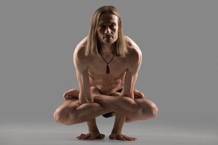 Sporty muscular young yogi man with long hair sitting in kukkutasana posture, doing cock pose, studio shot on dark grey background, front view, full lengthの写真素材