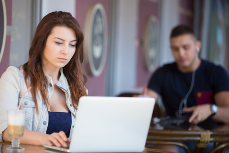 Portrait of serious good looking young woman sitting in street cafe with glass of drink on the table, working on laptop, looking at the screen, male customer on the backgroundの写真素材