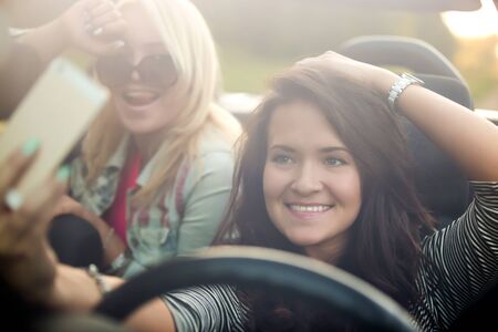 Two positive beautiful traveler young women taking selfie portrait with mobile phone camera in car, smiling, posing, having fun togetherの写真素材
