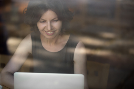 Portrait of happy smiling beautiful young woman sitting in front of window, using laptop, looking at the screen, female model working, interior shot through the glassの写真素材