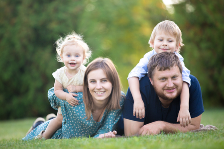 Mother, father and two kids lying on grass in park. Portrait of happy beautiful family of four relaxing on meadow, having fun together, spending leisure time on picnic in summerの写真素材
