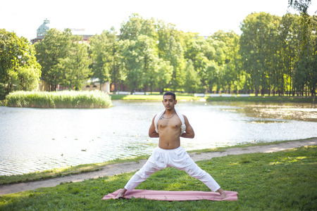 Attractive Indian young man in white linen clothes working out on river bank in park, doing preparation for Pyramid Pose, Intense Side Stretch Posture, parsvotanasana, hands behind back, full lengthの写真素材