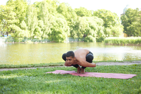 Serene attractive sporty Indian young yogi man working out on river bank in park, doing yoga, fitness or pilates training, sitting in Garland Pose, Malasana, full lengthの写真素材