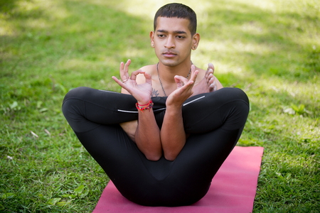 Attractive Indian young man in black sport leggings practicing yoga, fitness or pilates on red mat in park, doing garbhasana - fetus posture with hands in Namaste, front viewの写真素材