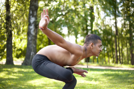 Sporty handsome Indian young man working out in park, preparing for Half Lotus Tip Toe Pose, Ardha Baddha Padma Padangusthasana, Padangustha Padma Utkatasana, side viewの写真素材