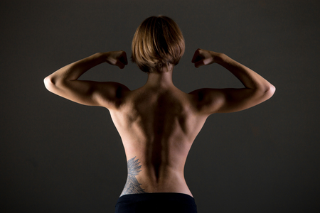 Young slim beautiful woman, with perfect body working out, posing, showing back muscles and biceps, body sculpture concept, studio image on grey backgroundの写真素材