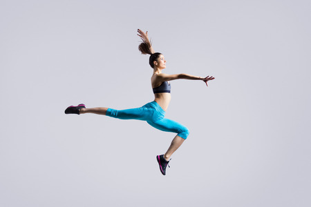 One attractive gorgeous young fit modern woman in blue sportswear with ponytail warming up, working out, dancing, jumping, full length, studio image on gray backgroundの写真素材